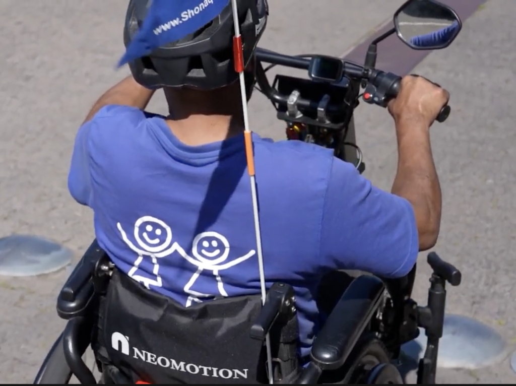 Rear view of a person riding a NeoMotion motorized wheelchair attachment wearing a Shonaquip Social Enterprise t-shirt